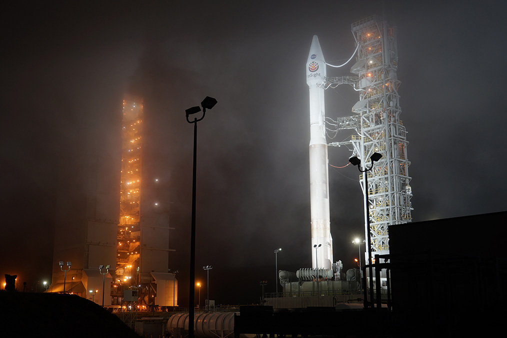  The mobile service tower shines brightly in the Californian night just a few hours before the final countdown. The Atlas V launcher is not yet visible from this viewing angle (© IPGP/Philippe Labrot).  The reliable precision engineering of United Launch Alliance (ULA), the consortium formed by Lockheed Martin and Boeing, worked perfectly once again for the InSight mission. On 5 May 2018 at 04:05, an Atlas V launcher lifted off from Vandenberg AFB Space Launch Complex 3, carrying the InSight geophysics observatory and the two MarCo nanosatellites on the first leg of their journey to the Red Planet.  As the countdown neared zero, thousands of people all along the rocket’s West Coast trajectory held their breath. Many must have been disappointed. The fog covering Vandenberg was so thick when the first-stage engine was fired that the Atlas V launcher’s ascension was completely hidden from view. Only an increasingly powerful roar resounding downwards in waves attested that the launcher was in fact climbing steadily upwards.  Despite the fact that practically nothing could be seen, everything went according to plan. The successful launch of InSight was not only a huge reward and relief for all the teams involved in the mission, but a tremendous testament to the ingenuity and dedication of the men and women working for Lockheed Martin, Boeing, the US Air Force and NASA, who are responsible for the launch.  The Atlas V launcher While the launch itself lasts less than two hours, it takes United Launch Alliance about one year to manufacture the various launcher parts, i.e. the central booster forming the first stage, the Centaur upper stage, the fairing and the many ring-shaped adapters that act as an interface between the different components. Most are made in plants on US soil, although the RD-180 engine that powers the first stage is actually Russian. Launch operations take two months. They consist in building the rocket on the launch pad like a giant Meccano® structure, and organizing everything to ensure a successful launch, from the availability of tracking facilities to the analysis of weather conditions.  A few hours before the launch, a visually impressive operation takes place. The mobile service tower used to work on the launcher is withdrawn. In the case of InSight, this operation was particularly spectacular due to the combination of fog and darkness. Looking like the kind of structure you’d find in an oil refinery, the tower was lit up like a Christmas tree with white and orange lights shining brightly in the Californian night. The rectangular edifice slowly rolled back along its rails, revealing the imposing Atlas V 401 launcher, whose pointed fairing proudly displayed the mission’s colours. Without its strap-on boosters, the rocket looked like a huge pencil pointing skywards, impatient to climb up into the night sky. Standing 57 metres tall, the launcher’s white cylindrical structure was lit up by the powerful beams of projectors that the US Air Force personnel changed from time to time to facilitate photography.  The beautiful yet intimidating complexity of the launcher and its support equipment awakens in onlookers the awareness that humans have an innate desire to explore. This positive, luminous aspect that probably resides in the innermost part of the human soul suddenly becomes crystal clear before such a scene. How else is it possible to explain why people devote so much time, effort, money and determination to deploy such sophisticated vehicles that, just a few minutes after their departure, are swallowed up in the bottomless abyss of space with no chance of returning, their destiny being to try to unveil the countless mysteries of the Universe in which we live?   The Atlas V launcher in all its splendour after the service tower’s rollback (© IPGP/Philippe Labrot).